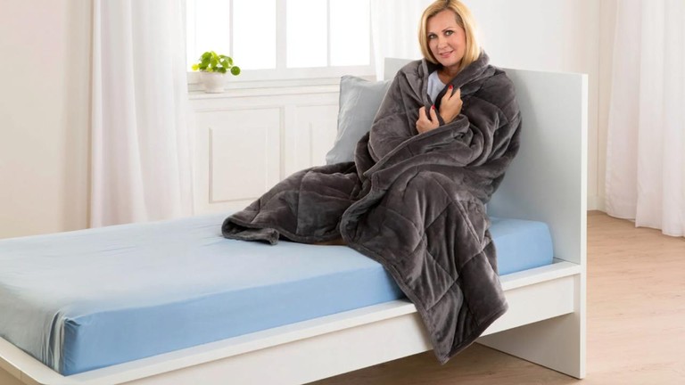 Terapeutická deka 8 kg ako upokojujúci darček na relax, prehodená cez ženu sediacu na posteli.